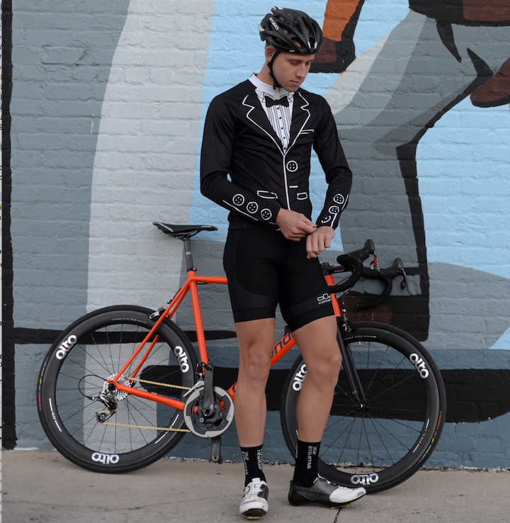 Rider in the Corbah Tuxedo long sleeve jersey standing with an orange road bike in front of a mural