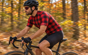 Side view of a cyclist riding through autumn woods in The Lumberjack flannel-print cycling jersey. Bib shorts sold separately.