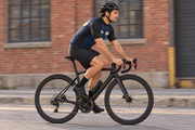Side view of a cyclist riding past a red brick building in The Mechanic navy jersey with charcoal sleeves. Bib shorts sold separately.
