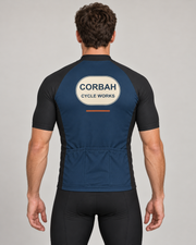 Back of The Mechanic jersey on a rider: navy body, charcoal raglan sleeves, CORBAH CYCLE WORKS badge with rust dash and three plain navy rear pockets. Bib shorts sold separately.