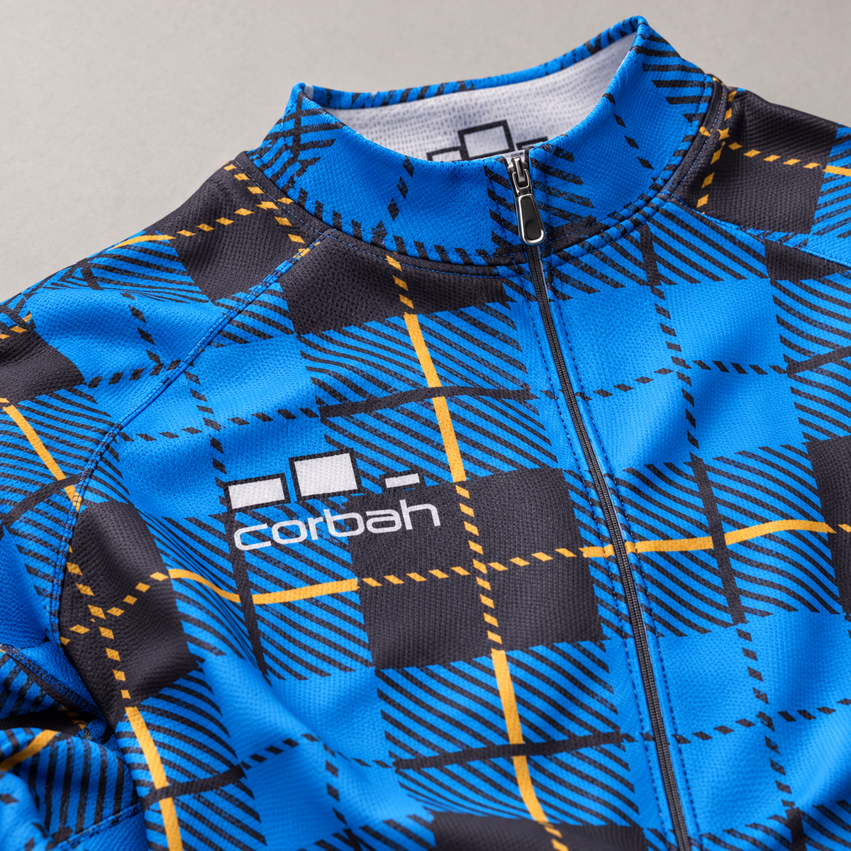 Close-up of the Scotsman jersey chest: white Corbah logo, front zip, collar and hatched blue, black and gold plaid fabric