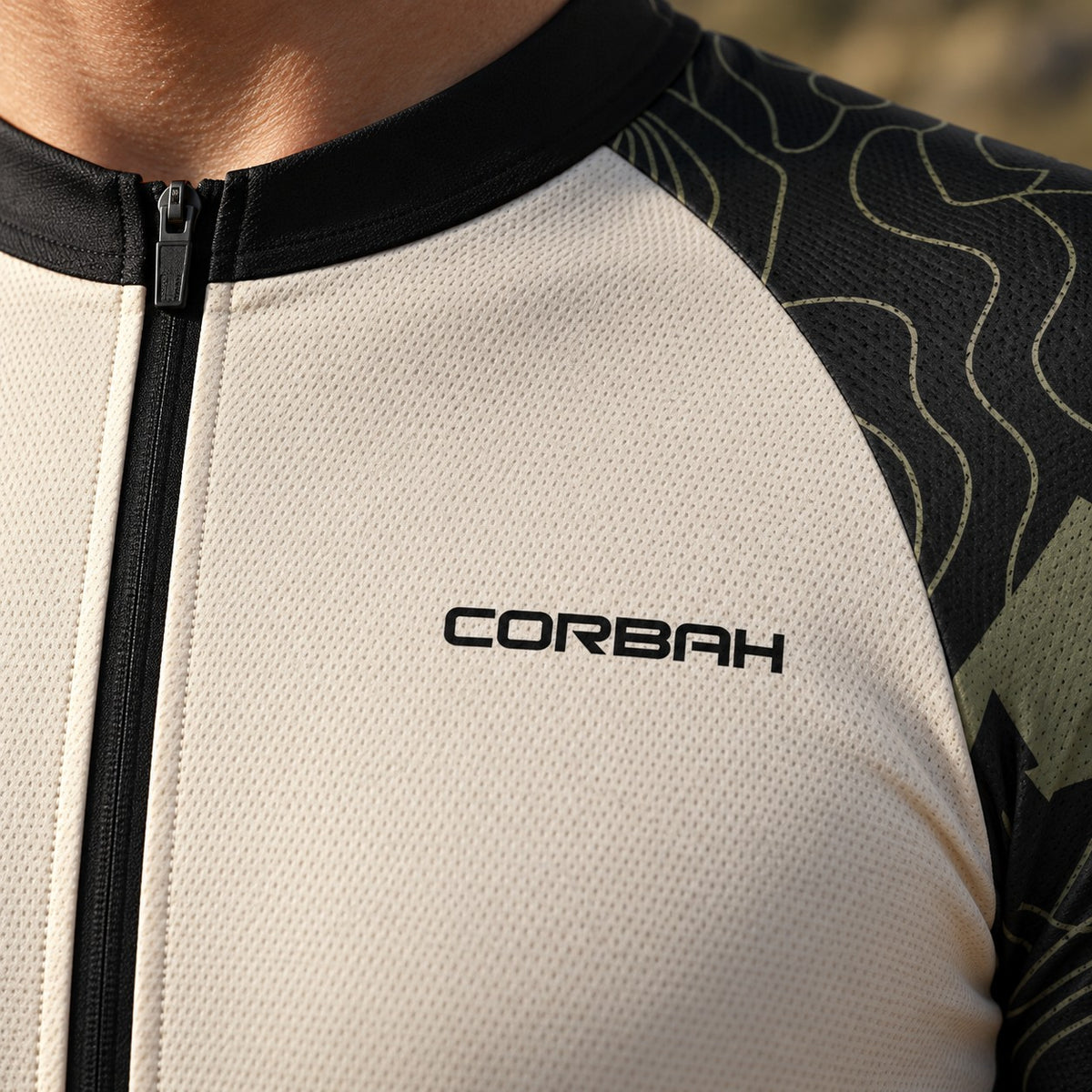 Close-up of the Corbah TOUGH jersey chest: black Corbah wordmark on bone fabric, black collar and zip, and olive topographic lines on the charcoal sleeve (AI visualization).