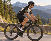 Side view of a rider in the Corbah TOUGH jersey climbing a mountain road past pines and granite, showing charcoal topo side panel, olive pine ridge and rust cuff bars on the sleeve (AI visualization; bib shorts not included).